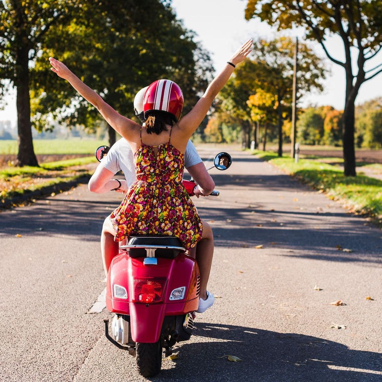 Zwei Leute fahren auf einem Roller in sonniger Umgebung, die Frau hat die Arme ausgebreitet.