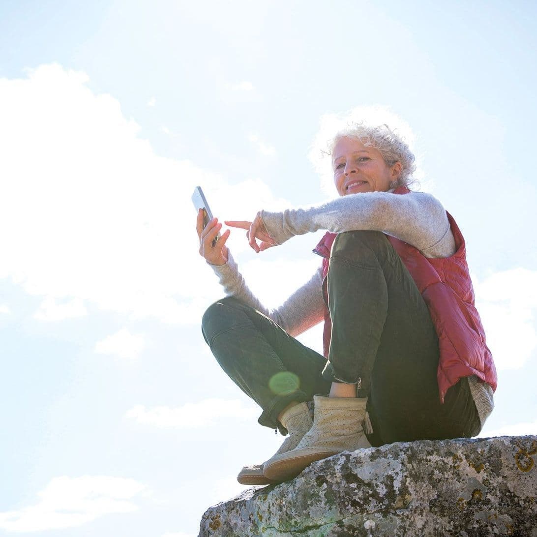 Ältere Frau sitzt auf einem Felsen und hält ein Handy in der Hand.